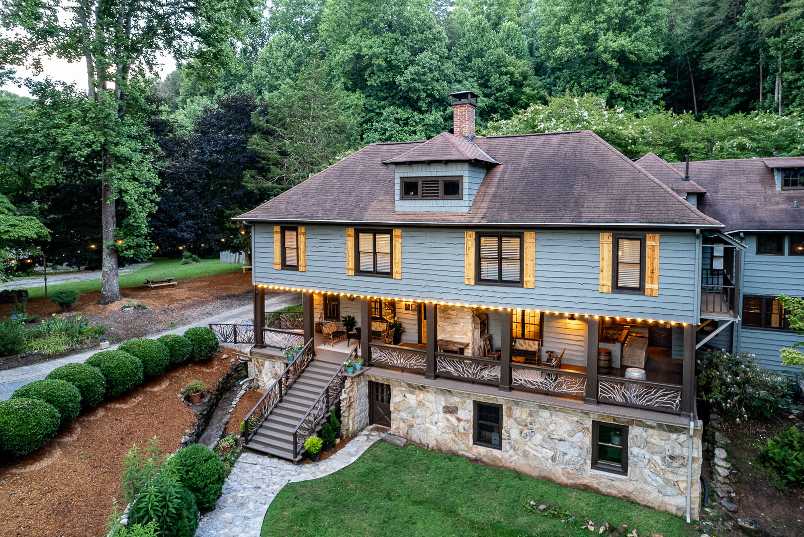 Blue wooden Bed & Breakfast with a stone foundation and wrap-around porch, lit by string lights in a wooded yard. - Clayton, GA
