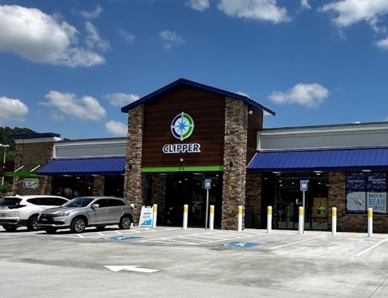 Exterior of a Clipper store with stone pillars, brown wood facade, blue awnings, and parked cars under a sunny sky. - Clayton, GA