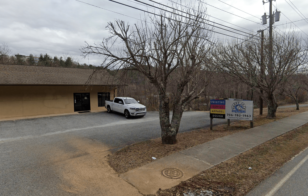 A small business storefront with a gravel parking lot, a white pickup truck parked near the entrance, and bare trees in front; a sidewalk runs along the street on the right under a cloudy sky. - Clayton, GA