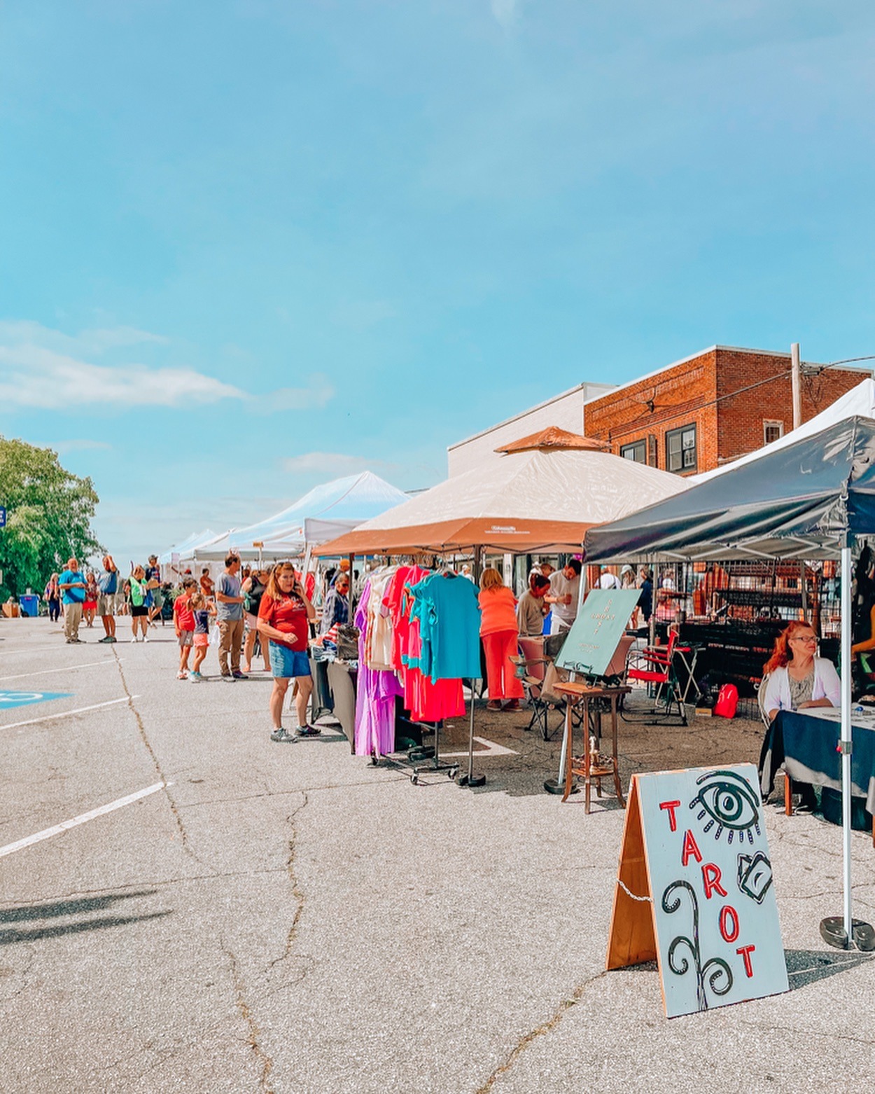 Outdoor street market with tents and shoppers browsing bright clothing on racks and tables under clear blue sky. - Clayton, GA