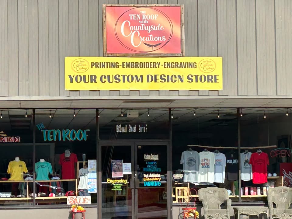 The Ten Roof With Countryside Creations - Visit Clayton, GA Front of a custom design shop with a bright yellow sign reading 'Your Custom Design Store' and t-shirt displays in the window. - Clayton, GA