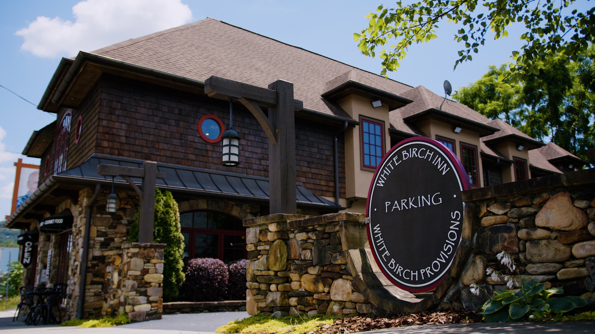 Exterior view of a rustic inn with brown shingles, stone wall, and hanging lanterns; a large circular sign reads 'White Birch Inn Parking' in the foreground. - Clayton, GA