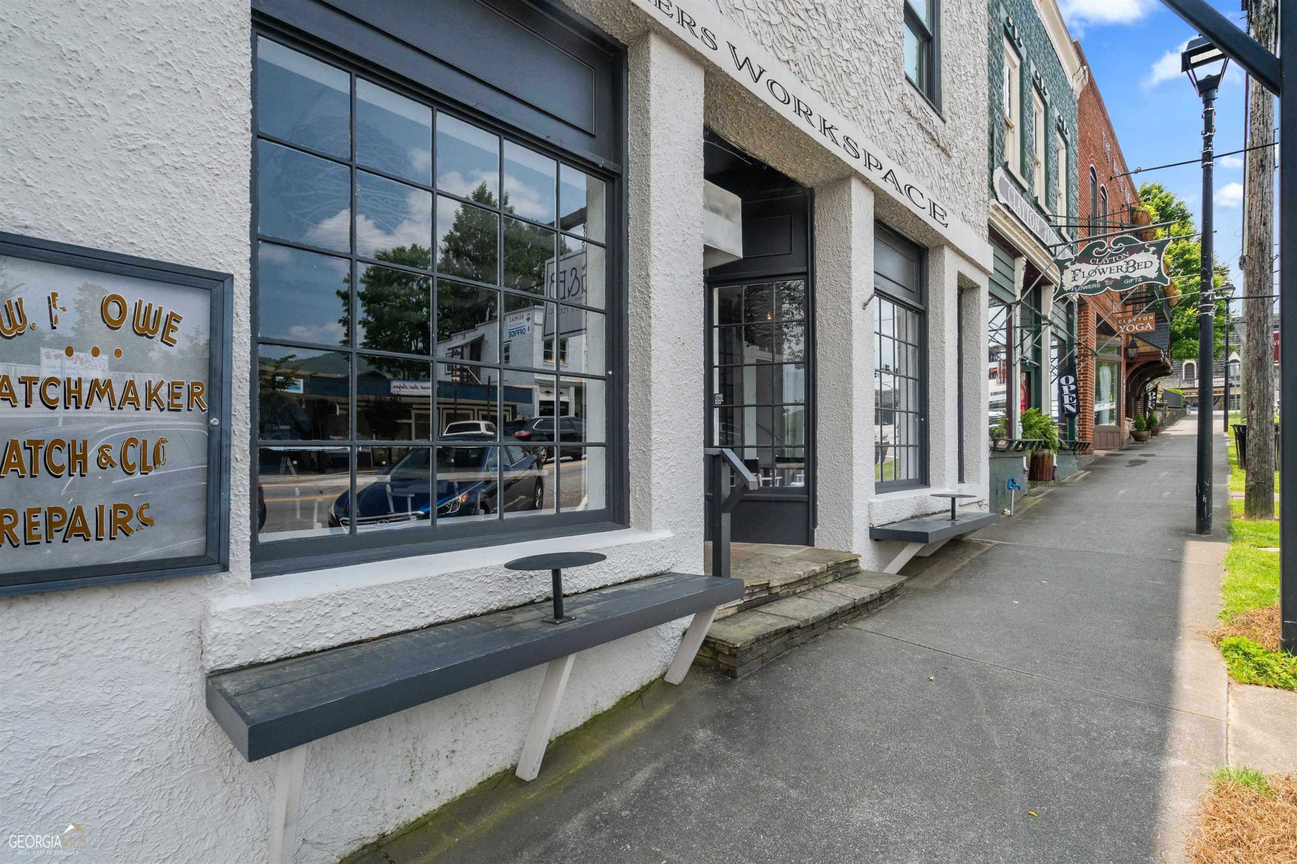 Row of storefronts along a sunny street with large grid windows, white textured walls, and a Clayton FlowerBed Gifts sign nearby indicating a flower shop. - Clayton, GA