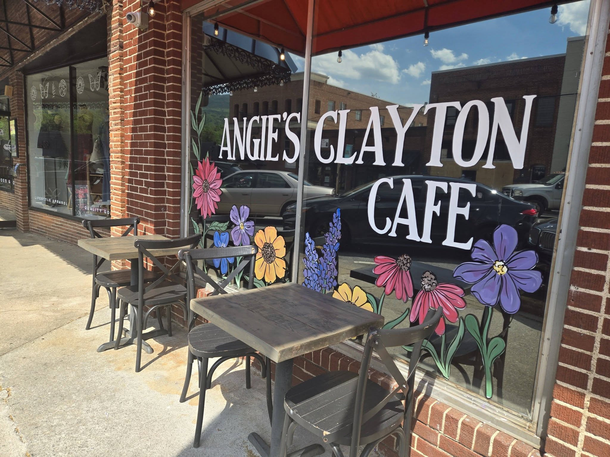 Storefront window with large white lettering and bright flower illustrations; small outdoor tables and chairs sit on the sidewalk beside a brick building. - Clayton, GA