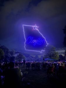 Nighttime outdoor event with a large dotted LED square frame and a blue glow in the sky above a seated audience in a park.