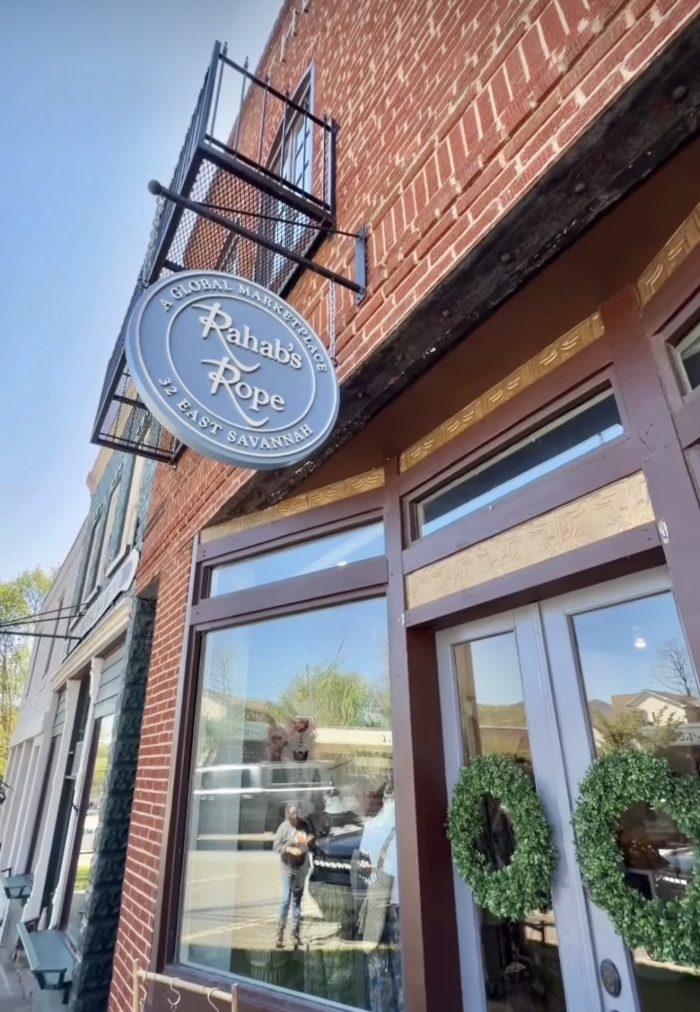 Circular blue sign reading 'Rahab's Rope' with 'A Global Marketplace' and '32 East Savannah' on a brick storefront exterior.