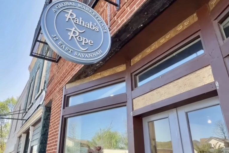 Circular blue sign reading 'Rahab's Rope' with 'A Global Marketplace' and '32 East Savannah' on a brick storefront exterior.