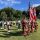 Flag party and musicians in kilts and red coats stand in a grassy field during an outdoor ceremony, holding US and Canadian flags and instruments nearby.