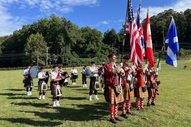 Flag party and musicians in kilts and red coats stand in a grassy field during an outdoor ceremony, holding US and Canadian flags and instruments nearby.