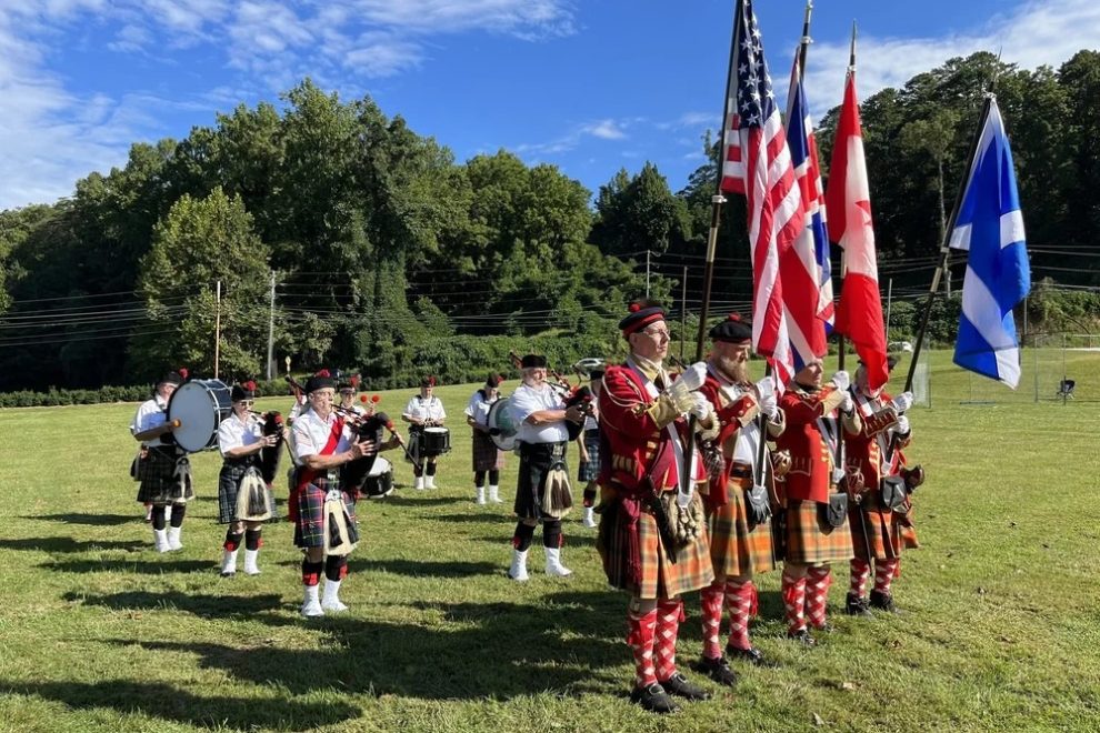 Flag party and musicians in kilts and red coats stand in a grassy field during an outdoor ceremony, holding US and Canadian flags and instruments nearby.