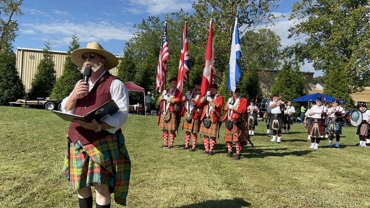 Man wearing a straw hat and kilt speaks into a microphone while leading a bagpipe band outdoors, with several flag bearers in the background.