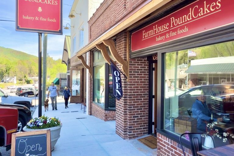 Exterior view of FarmHouse PoundCakes bakery on a sunny street, brick storefront, red sign, and open doorway with a chalkboard menu outside.