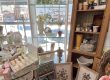 Interior of a boutique with a tall white wicker arched display shelf holding mugs and glassware, in front of a window.