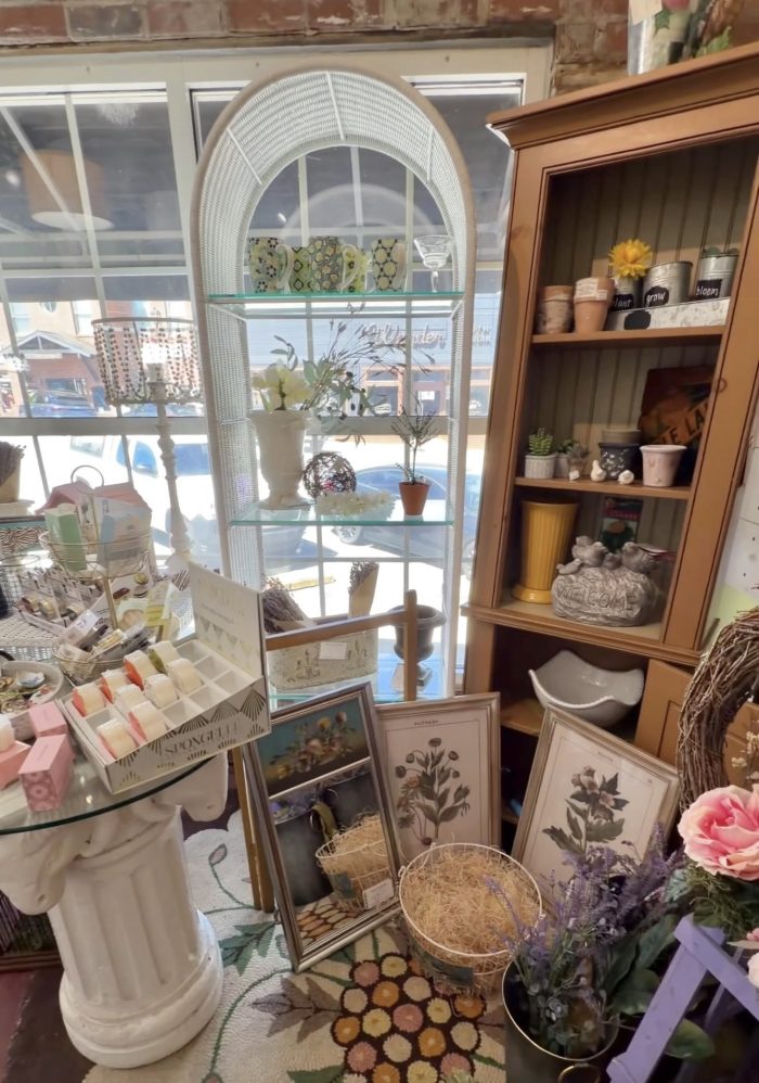 Interior of a boutique with a tall white wicker arched display shelf holding mugs and glassware, in front of a window.
