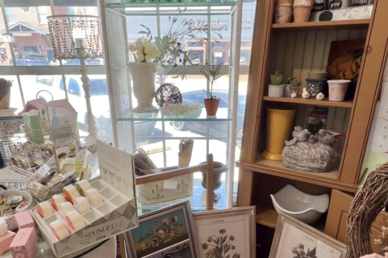 Interior of a boutique with a tall white wicker arched display shelf holding mugs and glassware, in front of a window.