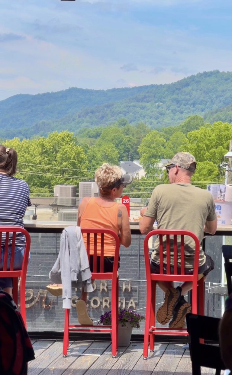 People sit at a railing-view outdoor cafe with bright red chairs, backs to camera, overlooking green hills and mountains.