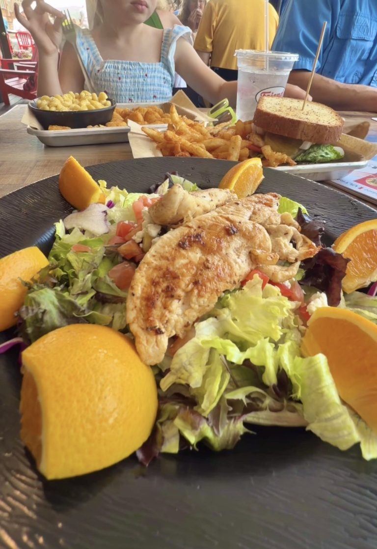 Plate with grilled chicken on a mixed greens salad and orange wedges; to the side are breaded fried items and fries, with a sandwich and drink in the background.