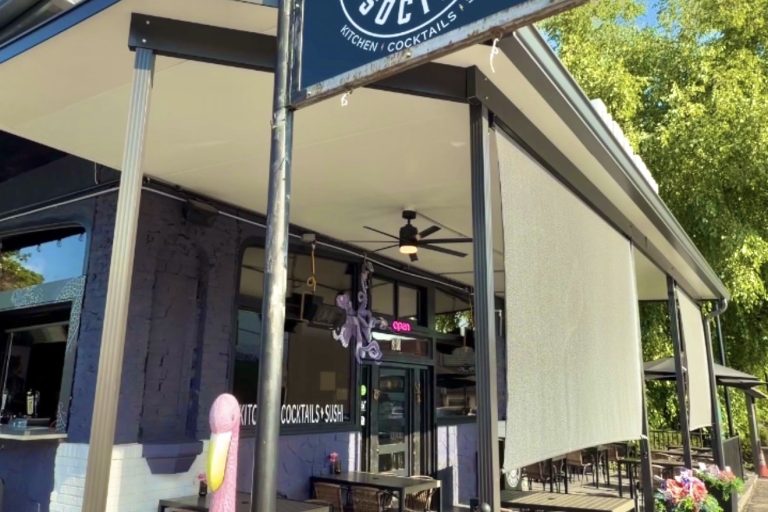 Exterior view of Rabun Social restaurant with a large black sign showing a tiger wearing sunglasses and the text ‘Kitchen Cocktails Sushi,’ outdoor seating under a covered patio.