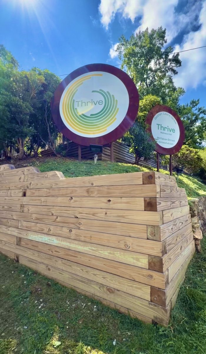 Circular sign with Thrive logo and green/yellow concentric rings, set on a wooden frame outdoors at Thrive Wellness Hub.