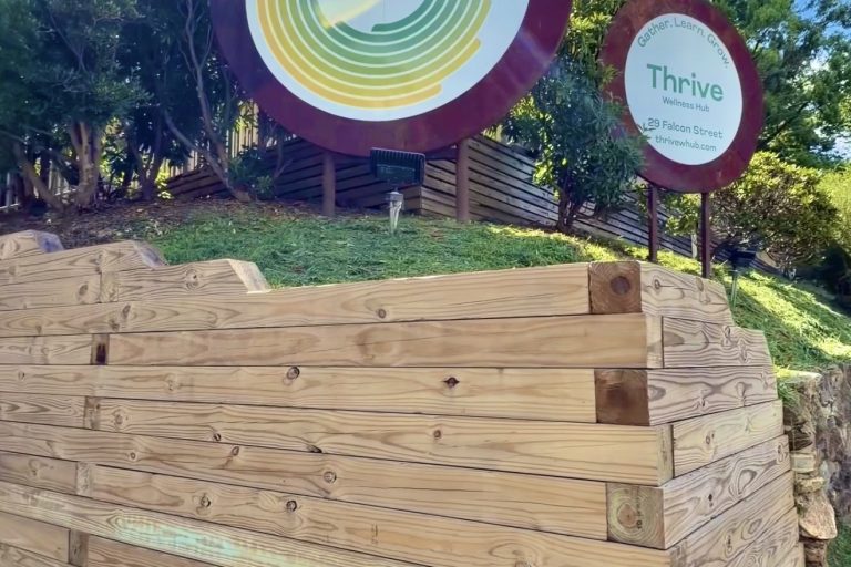 Circular sign with Thrive logo and green/yellow concentric rings, set on a wooden frame outdoors at Thrive Wellness Hub.