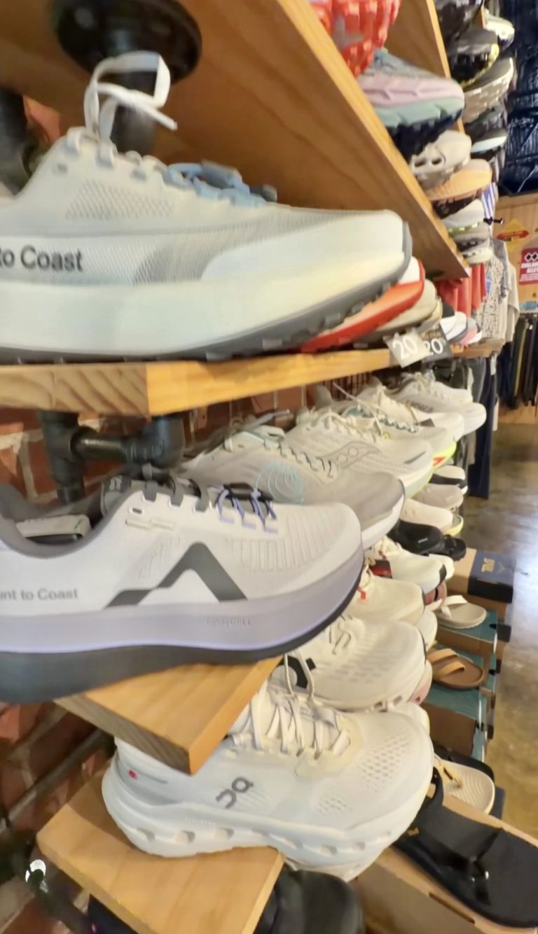 Light gray running shoes on a multi-tier wooden shoe display in a store, with many other sneakers lined up along the shelves