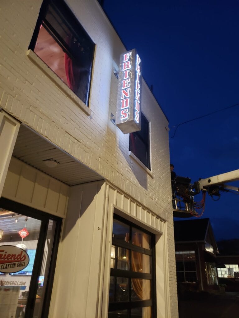 Friends American Grill - Visit Clayton, GA Nighttime exterior of a white brick restaurant with a vertical neon sign reading 'FRIENDS' on the corner. - Clayton, GA
