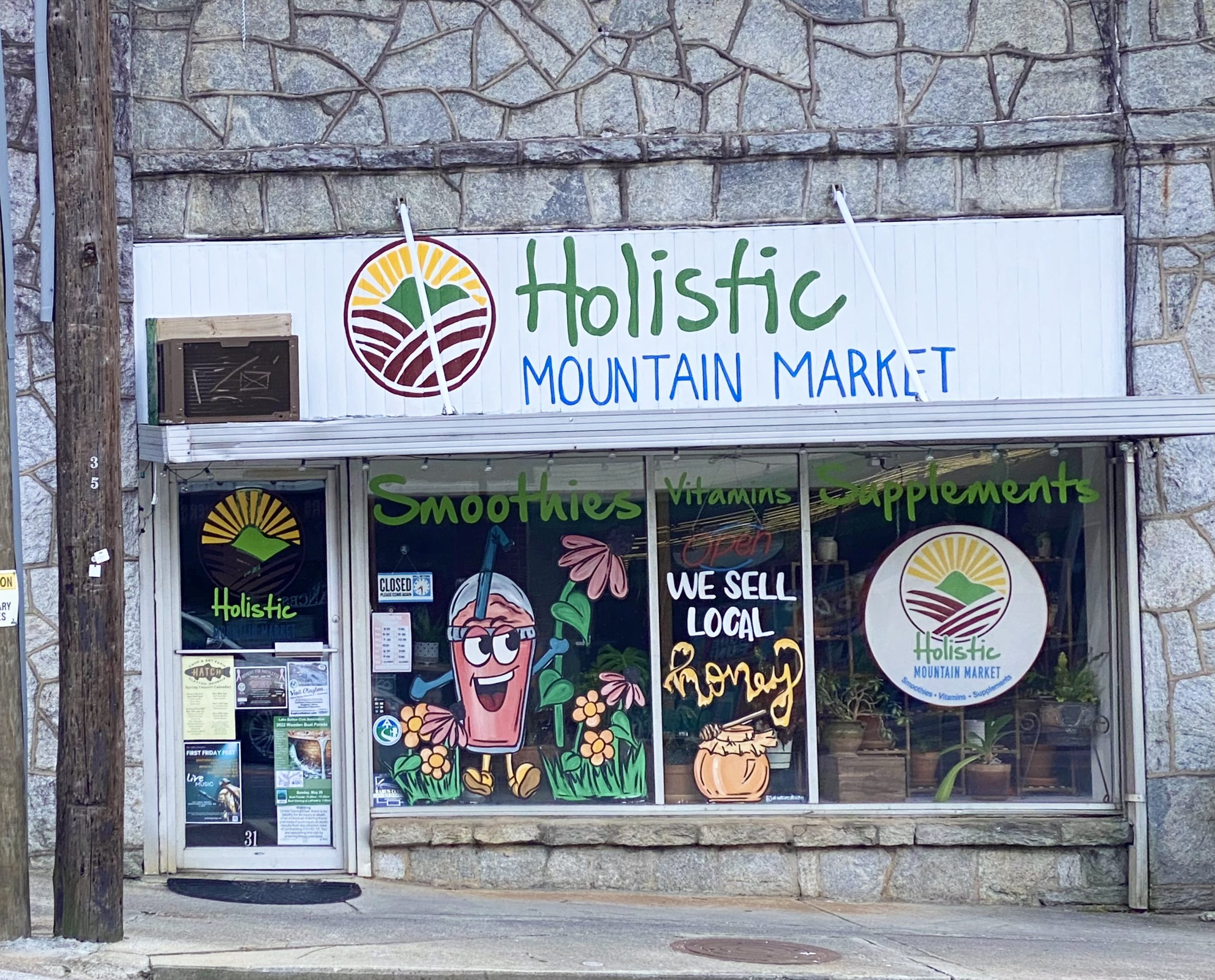 Holistic Mountain Market - Visit Clayton, GA Storefront sign for Holistic Mountain Market with a sun-mountain logo and colorful window art advertising smoothies and vitamins/supplements. - Clayton, GA