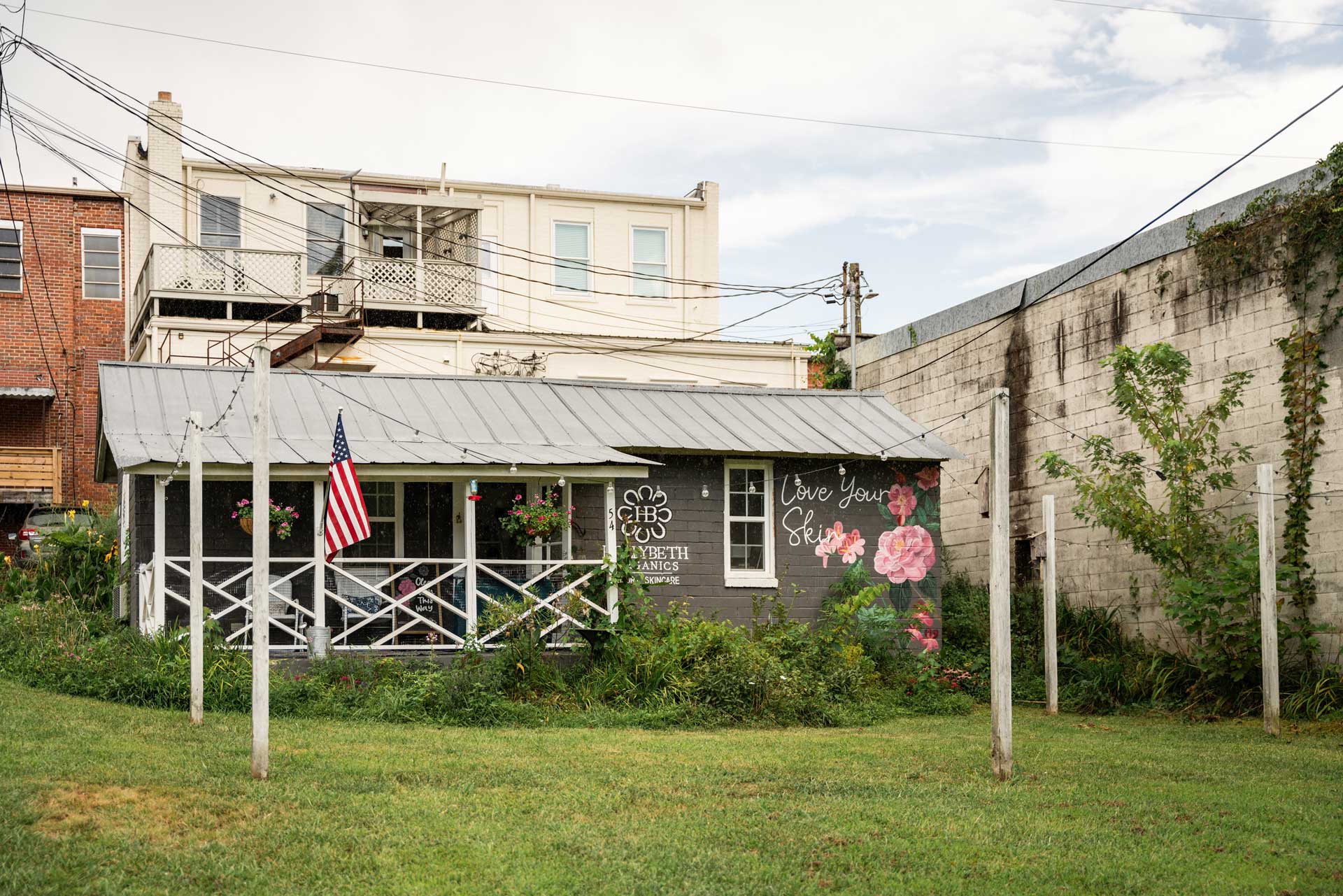 HollyBeth Organics - Visit Clayton, GA Small storefront with a dark gray wall featuring pink flower art and the words 'Love Your Skin'; an American flag on a pole stands in front under string lights. - Clayton, GA