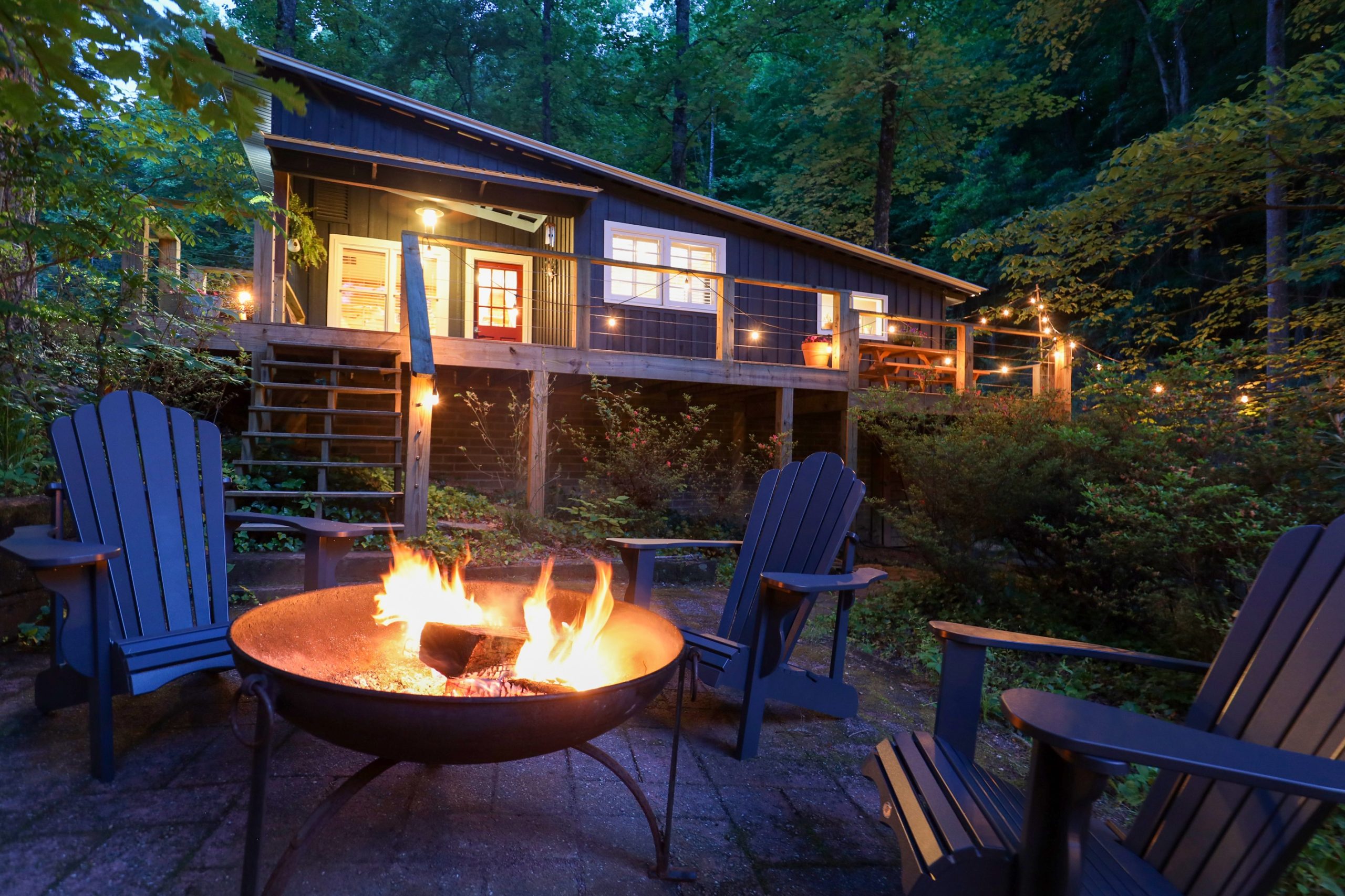 Just Right Vacation Rental Creekside Clayton - Visit Clayton, GA Nighttime backyard with a wooden cabin, string lights, and a glowing fire pit surrounded by blue Adirondack chairs. - Clayton, GA