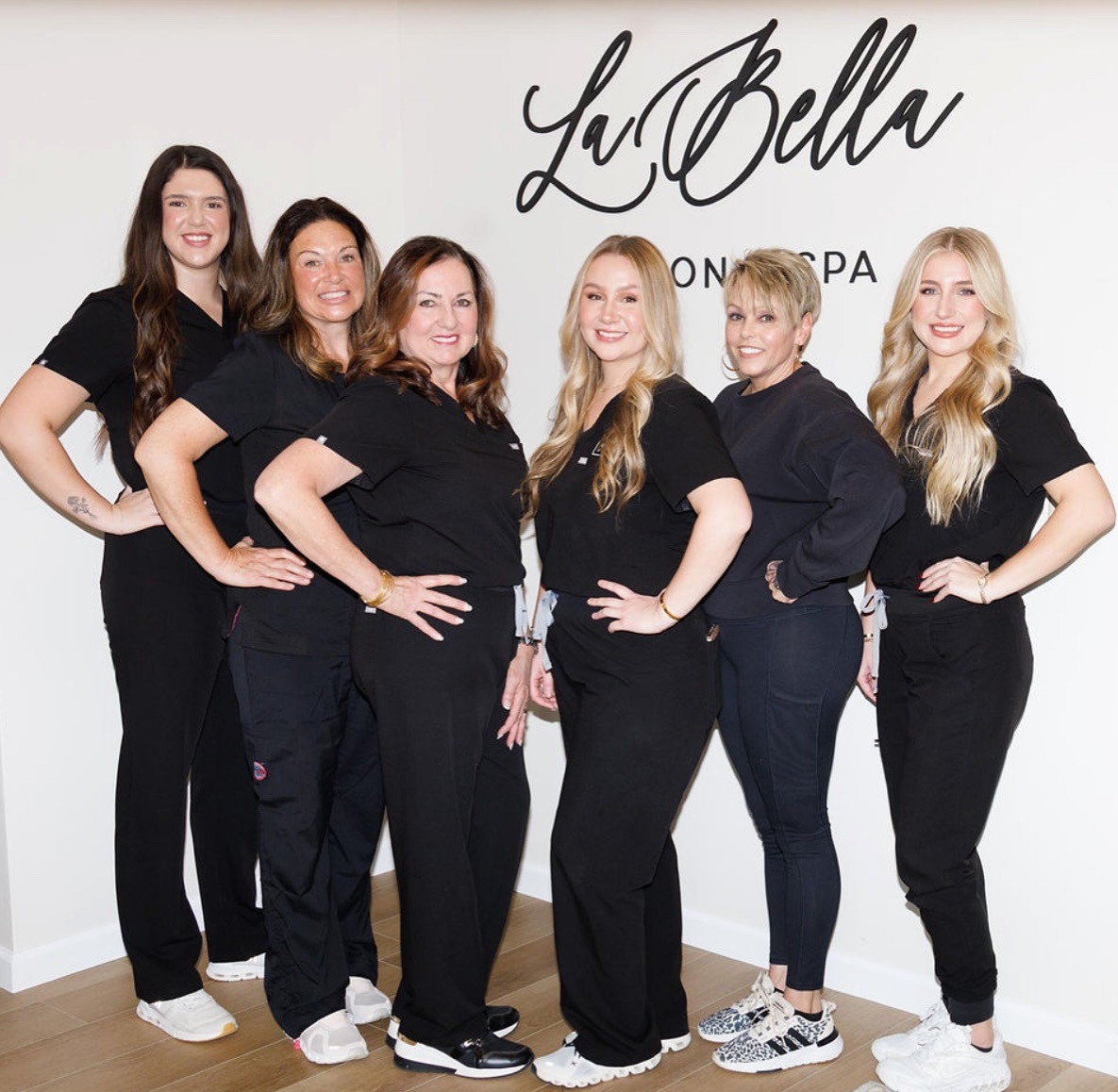La Bella Aesthetics - Visit Clayton, GA Six women in black scrubs pose together in a spa or salon, with the La Bella logo on the wall behind them. - Clayton, GA
