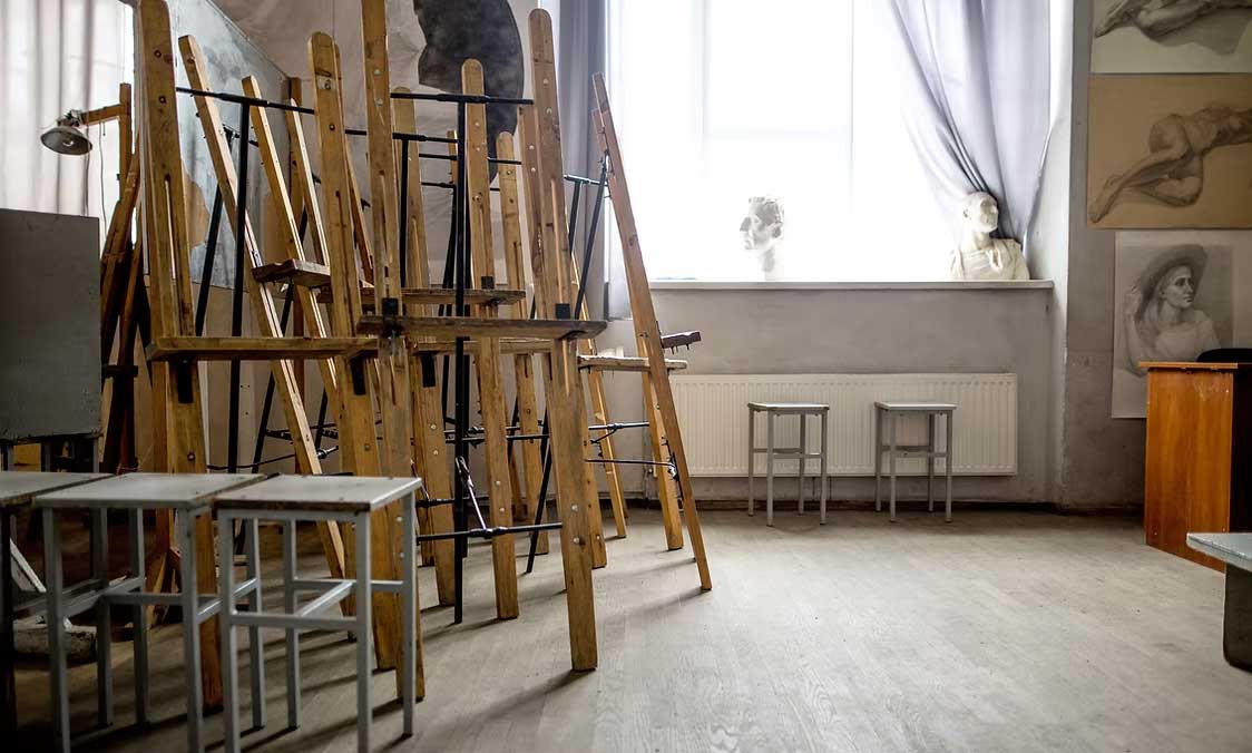 North Georgia Arts Guild - Visit Clayton, GA Art studio with tall wooden easels stacked together, metal stools, and sketches on the wall near a bright window with curtains. - Clayton, GA