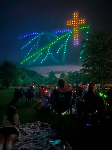 Outdoor nighttime scene: a crowd seated on blankets and chairs in a park, watching a colorful LED light installation forming a cross and palm-like rays in the sky.