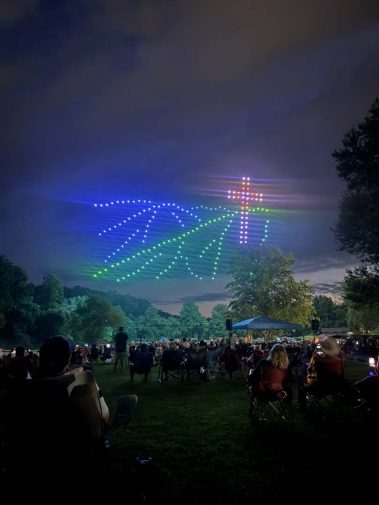 Crowd seated on a park lawn at night watching a rainbow LED light show in the sky at an outdoor event.
