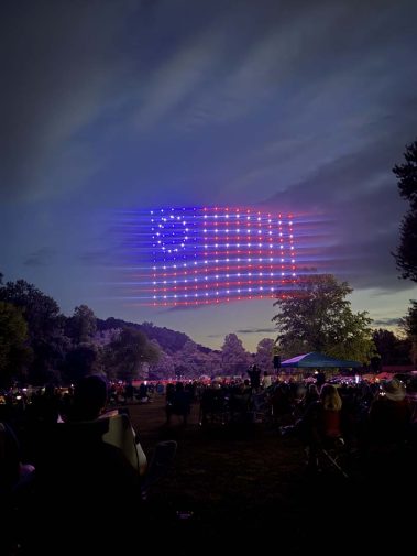 Evening outdoor event with a large LED grid forming an American flag in the sky, while people sit on the grass watching.