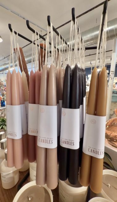 Tall tapered candles in neutral tones hanging from a metal display rack in a shop window.