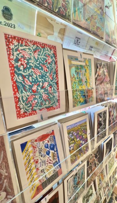 Row of colorful art prints displayed on glass shelves in a shop, featuring tropical motifs and bold red, teal, and yellow colors.