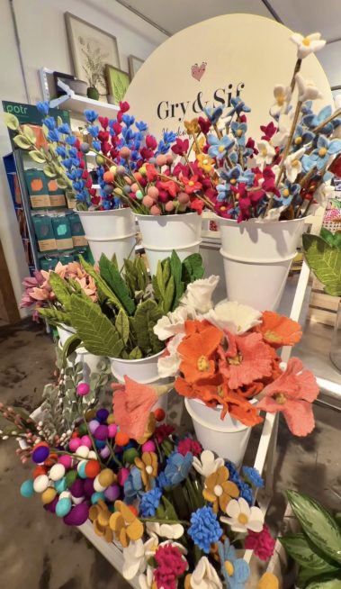 Display of colorful artificial flowers in white pots on a store shelf with a rounded sign in the background, bright blue, pink, and orange blooms present.
