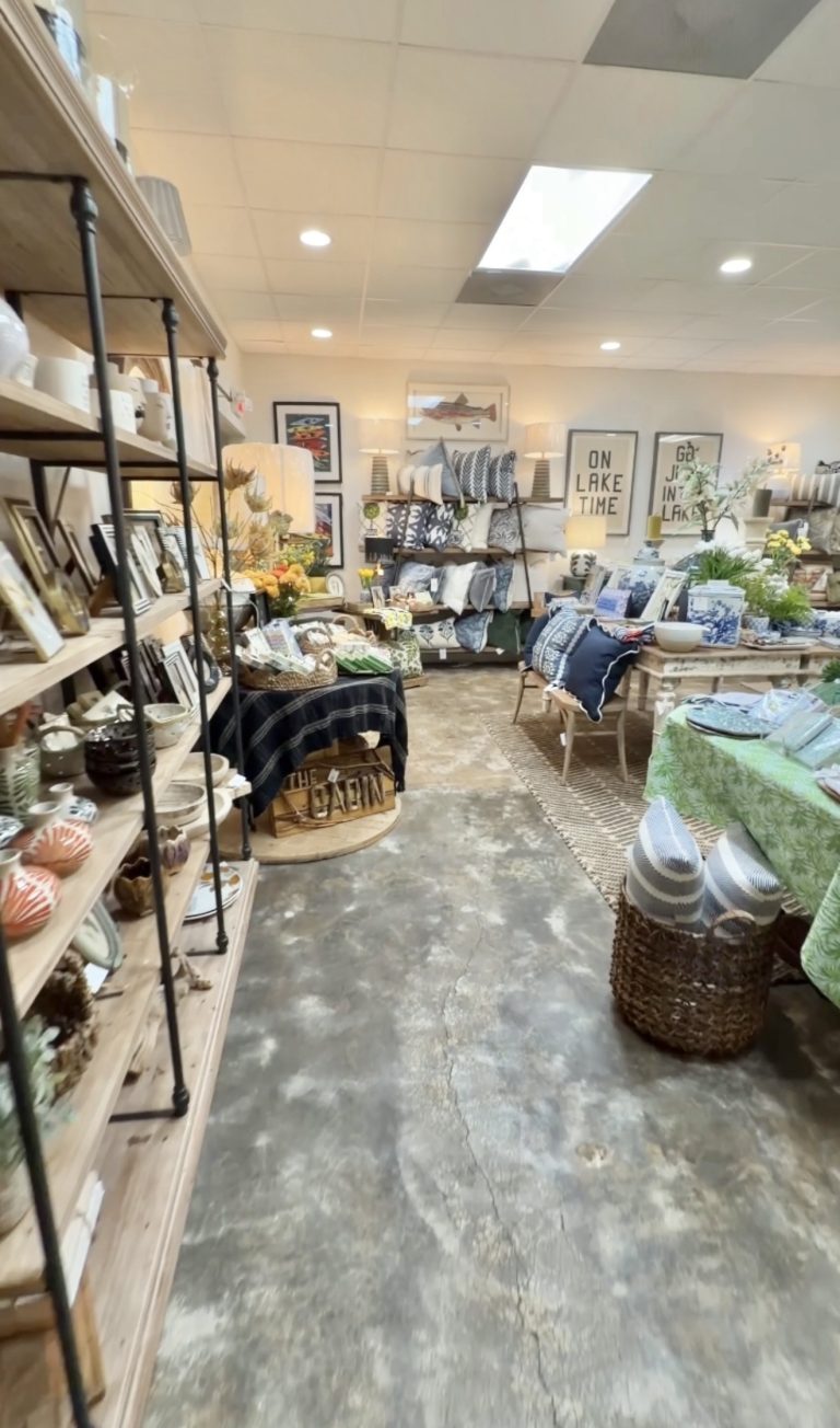 Interior of a home decor store with shelves of pottery and frames along a wooden display wall, and tables stacked with pillows, vases, and greenery.