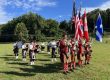 Flag party and musicians in kilts and red coats stand in a grassy field during an outdoor ceremony, holding US and Canadian flags and instruments nearby.