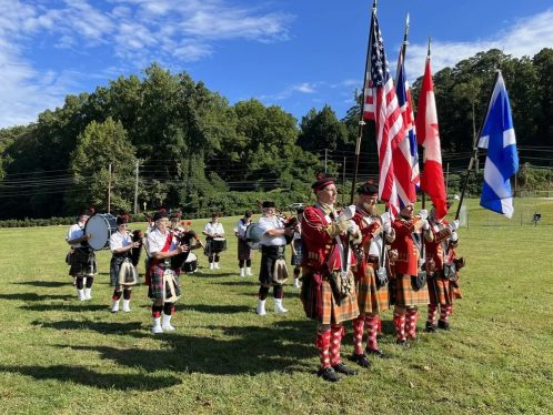 Flag party and musicians in kilts and red coats stand in a grassy field during an outdoor ceremony, holding US and Canadian flags and instruments nearby.