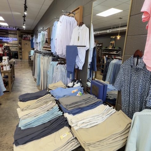 Men's and boys' dress shirts on racks with folded pants on tables in a clothing store showroom.