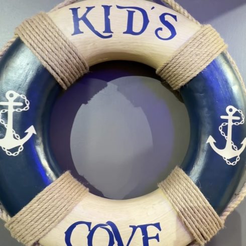 Decorative kids' lifebuoy with rope wrap, hanging on a wall; the word 'KIDS' is painted on the top segment and 'COVE' visible at the bottom.