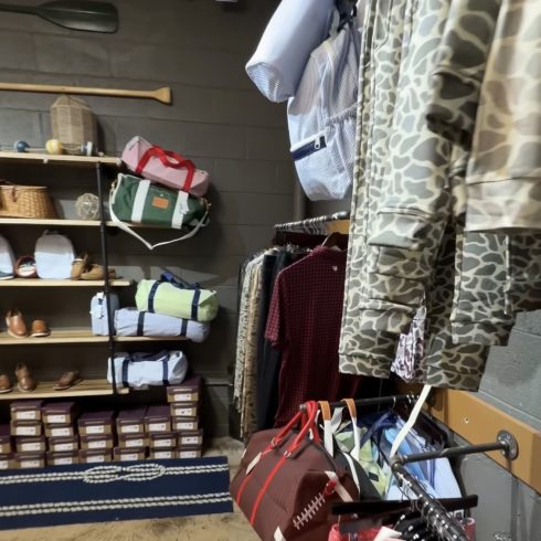 Clothes and bags displayed in a shop: camo jackets on racks, folded garments, and tote bags on shelves beneath a string of nautical flags.