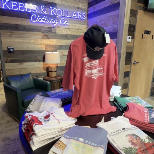 Display table with folded vintage-style t-shirts in front of a wood-paneled wall and neon sign reading 'KEELS & KOLLARS Clothing Co' behind a mannequin wearing a red shirt and a black cap.