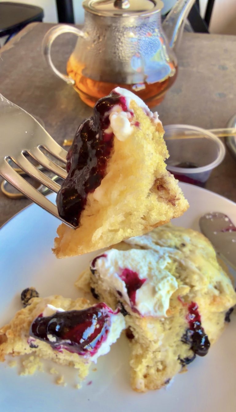 Close-up of a fork lifting a bite of yellow cake with blueberry jam and cream; plate with more cake and a glass teapot in the background.