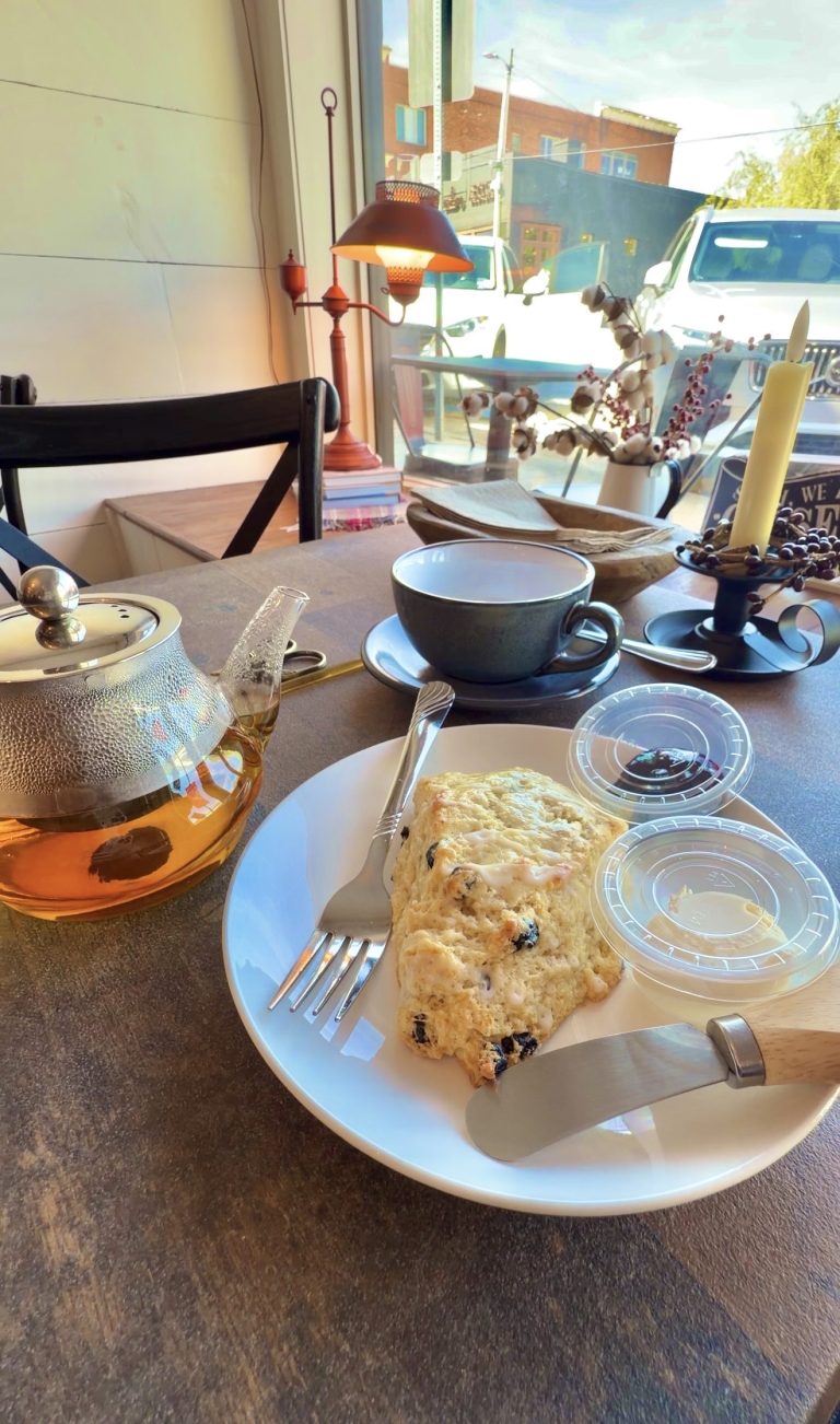 Plate with blueberry scone, fork, and clear plastic lid, alongside a glass teapot and blue teacup in a cozy cafe setting by a window.