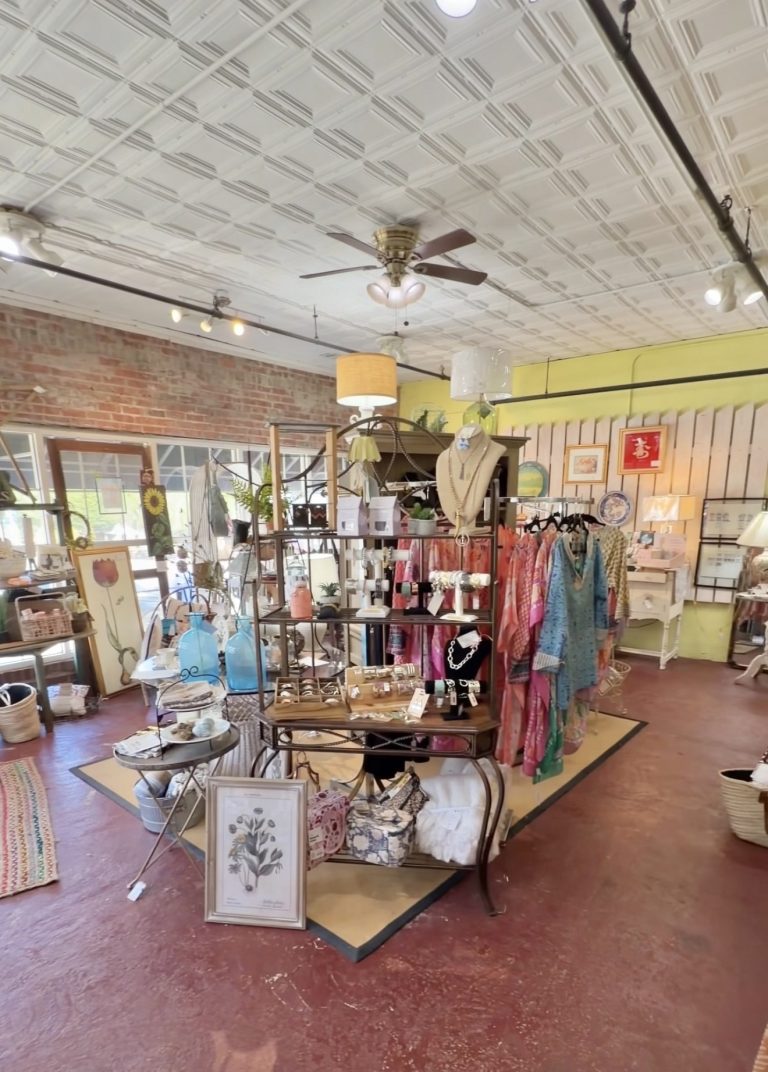 Boutique interior with brick walls, hanging clothing racks, and a central display table filled with accessories and framed art pieces under a patterned ceiling fan.