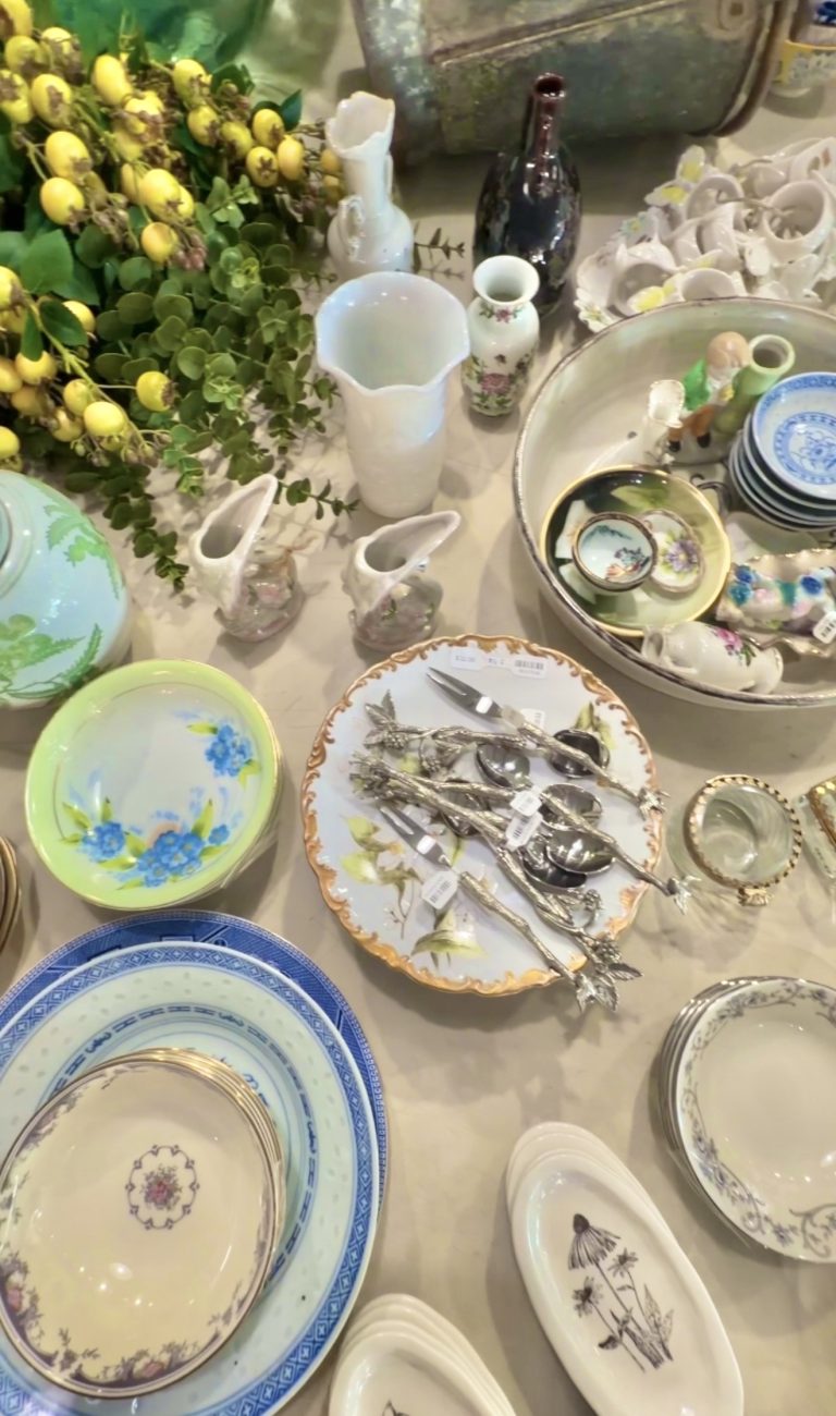 A cluttered display of ceramic dishware including blue and white plates, pastel bowls, and white decorative vases among yellow berry stems and greenery.