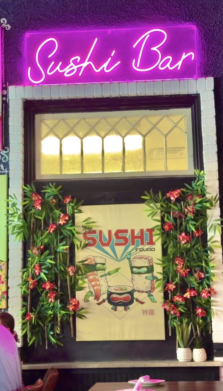 Bright pink neon sign reads 'Sushi Bar' above a window framed by tropical plants and pink flowers at the entrance.