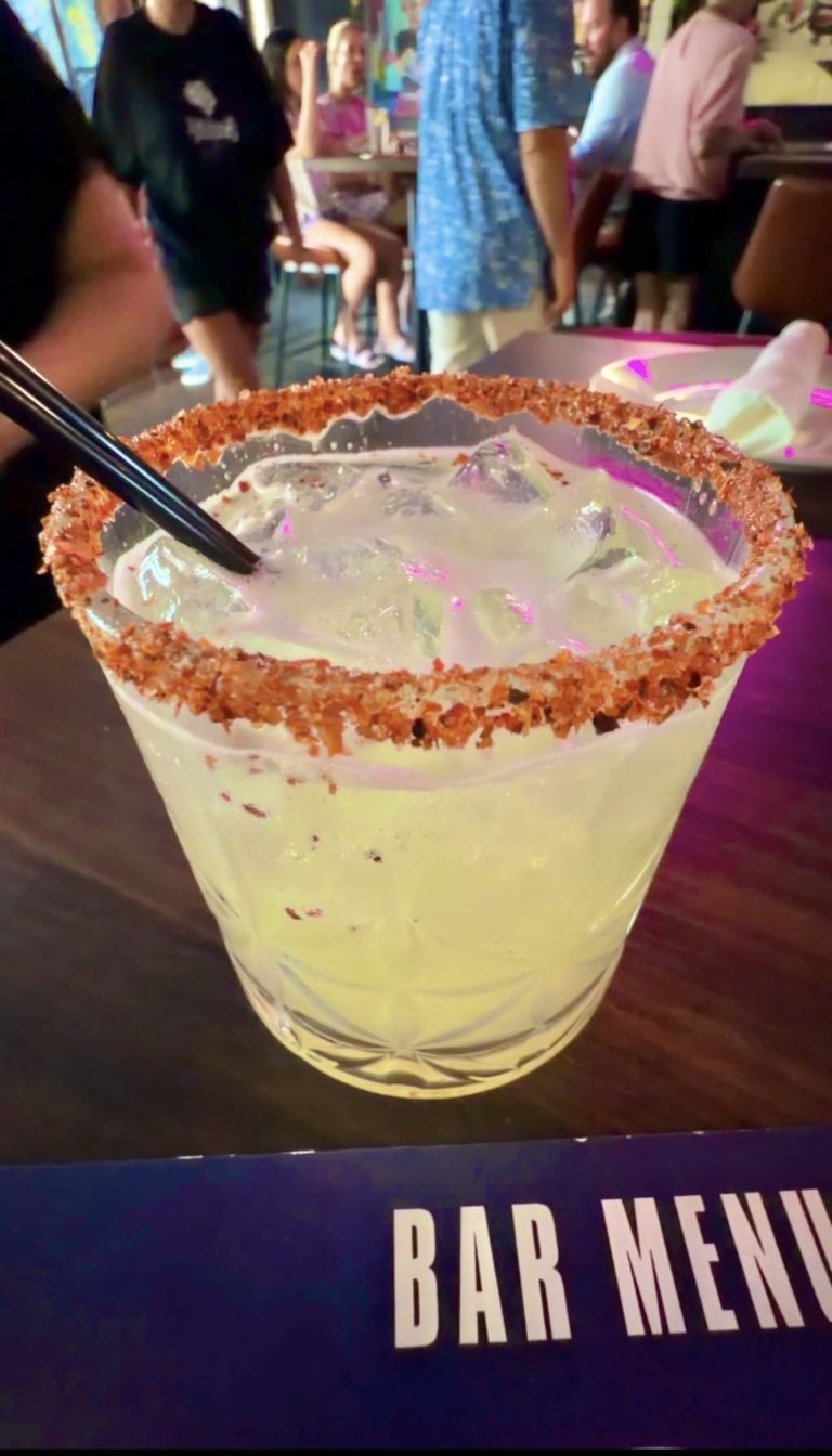 Close-up of a frosted margarita glass with a salted rim, blue-green icy drink, and a black straw, at a lively bar.